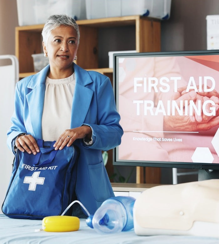 Woman holding a first aid kit in front of a screen displaying 'First Aid Training' in an indoor setting.
