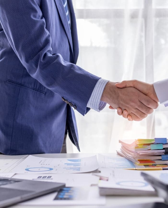 Two businesspeople shaking hands at a desk with financial documents and a first aid kits.