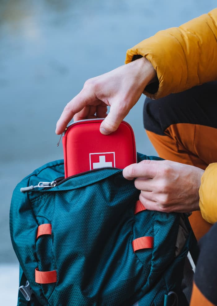 Person in a yellow jacket placing a red first aid kit into a teal backpack by a body of water.