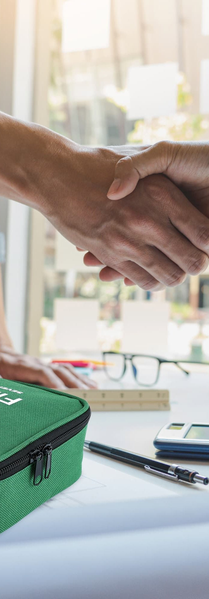 Two people shaking hands with a green first aid kit on a table in an office setting.